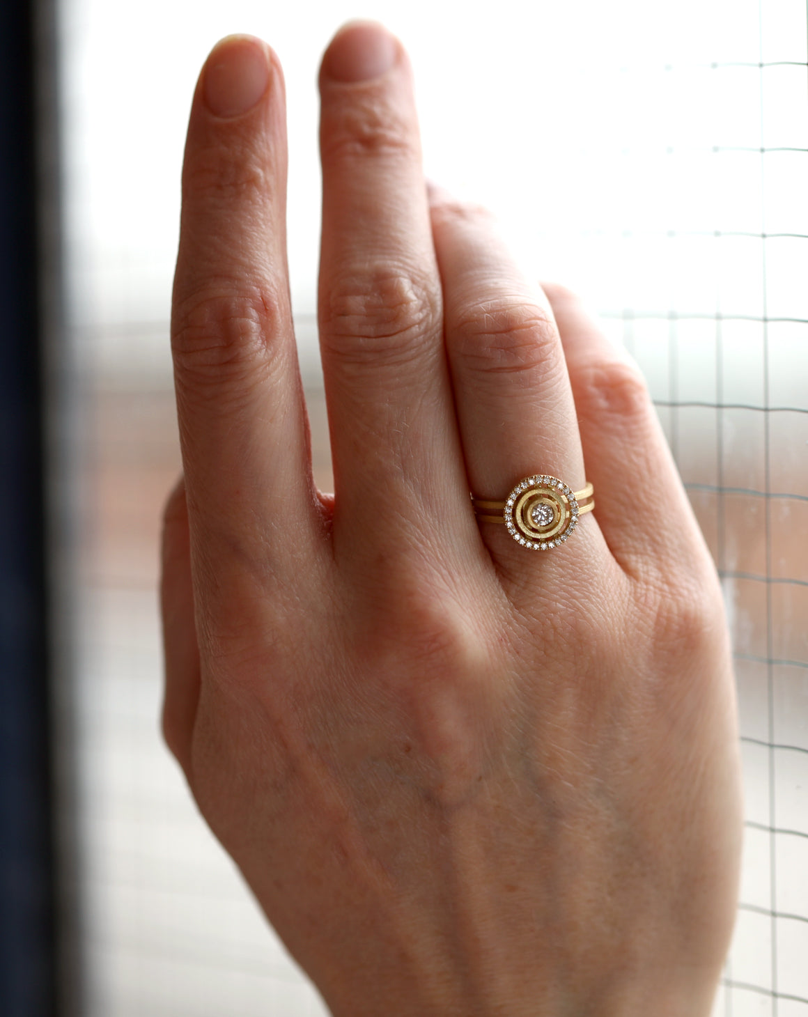 Geometric ring with concentric circular detail, diamond halo and central diamond. 18ct yellow gold textured finish. On hand.