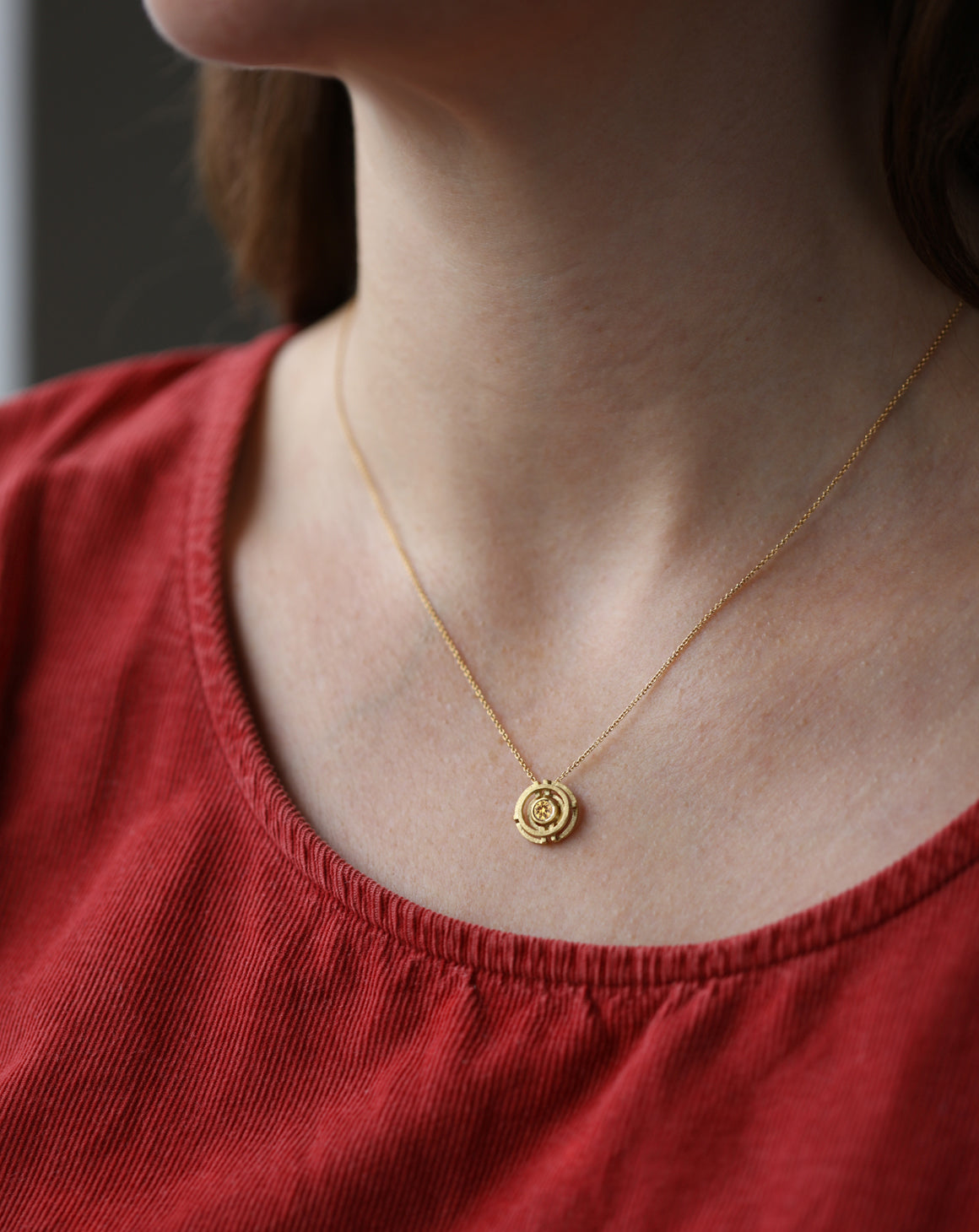 Geometric pendant with concentric circles surrounding yellow sapphire. 18ct yellow gold, textured finish. On neck.