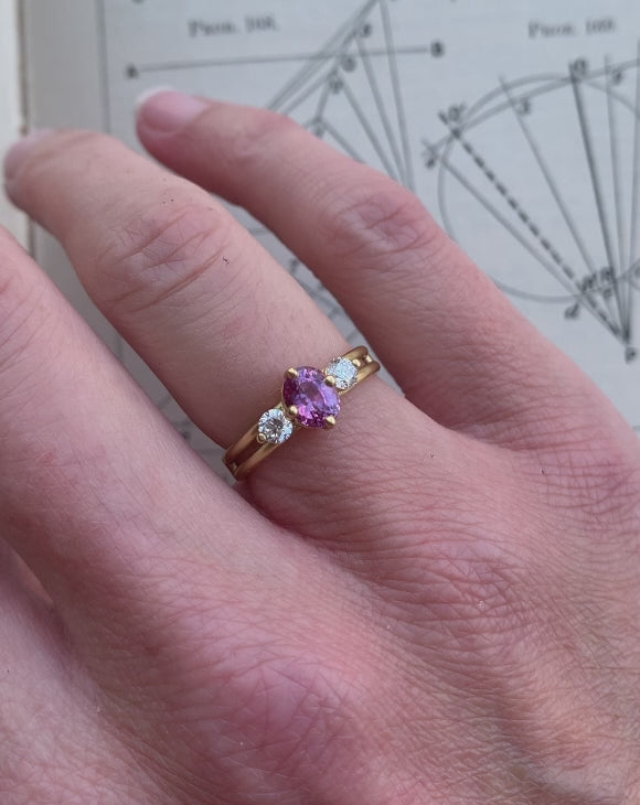 Three stone ring with a pink oval sapphire and two white diamonds. 18ct yellow gold, satin finish. On hand.
