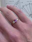 Three stone ring with a pink oval sapphire and two white diamonds. 18ct yellow gold, satin finish. On hand.