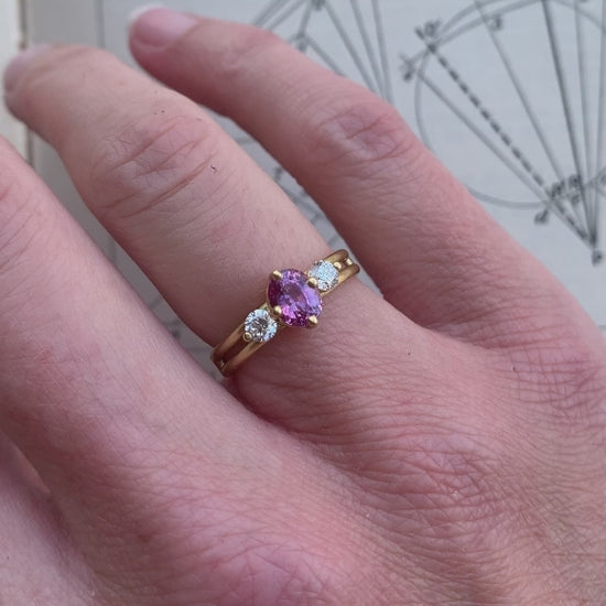 Three stone ring with a pink oval sapphire and two white diamonds. 18ct yellow gold, satin finish. On hand.